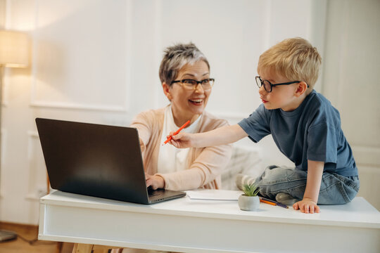 Senior woman with short gray hair and glasses interacts with young boy in blue shirt at a desk with a laptop and stationery in a bright, modern home office setting
