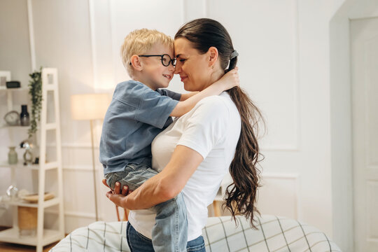 Caucasian woman holding young boy in blue shirt, both smiling and embracing in a bright, modern living room with soft furnishings and decorative shelves