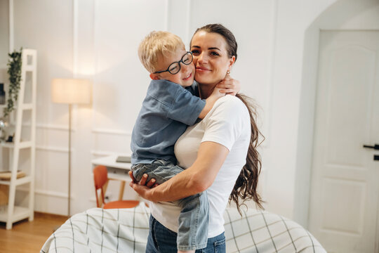 Caucasian woman holds young boy in her arms, both smiling, inside a bright living room with modern decor and a cozy sofa in the background
