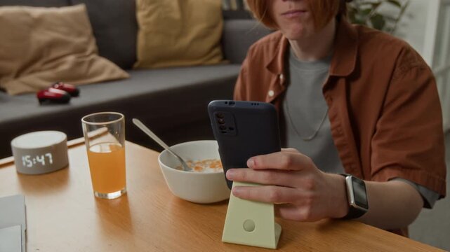 Teenage Caucasian boy wearing headphones eating breakfast while watching video on smartphone. Clip is perfect for promoting mobile apps, streaming services, or digital lifestyle brands