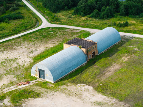 Aerial view of a metal Quonset hut hangar situated in a rural landscape with a dirt road and green fields in Kalnciems, Jelgava Municipality, Latvia.