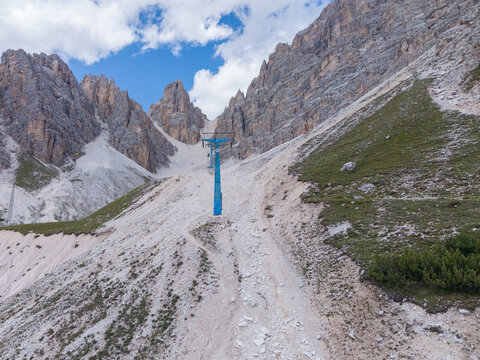 Aerial view of an abandoned chair lift tower standing on a rocky mountain slope with jagged peaks and sparse vegetation in Cortina d'Ampezzo, Veneto, Italy.