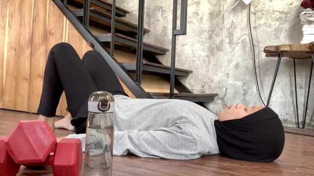 Slow motion shot of a young Muslim woman in a hijab doing a glute bridge exercise on a wooden floor at home. Concept of healthy active lifestyle and indoor fitness routine.