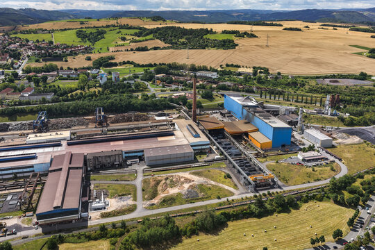 Aerial view of the industrial steel plant with large factory buildings, a tall chimney, and scrap metal piles surrounded by green fields and rolling hills in Unterwellenborn, Thuringia, Germany.