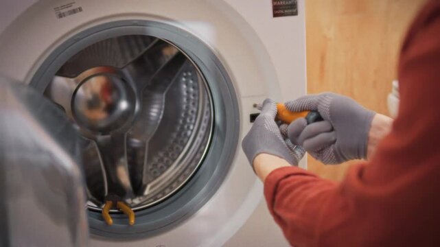A close-up of a technician's gloved hands unscrewing a bolt from the body of a washing machine in a bathroom with a toolbox in the background. Concept of washing machine maintenance and repair
