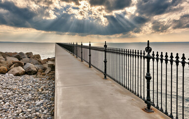 Obraz premium Concrete walkway along shingle beach next to ocean water. Black metal railing lines paved path towards horizon under cloudy sky.