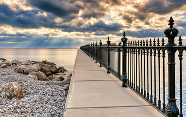 Obraz premium Concrete walkway along shingle beach next to ocean water. Black metal railing lines paved path towards horizon under cloudy sky.