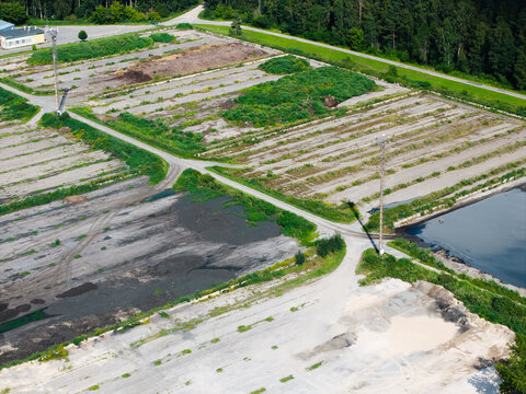 Aerial view of the wastewater sludge storage facility featuring multiple enclosed storage basins, reservoirs, and dirt access paths surrounded by dense forest in Riga, Latvia.
