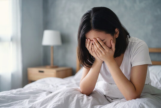 Depressed and sad young Asian woman sitting on bed and covering face with hands in bedroom