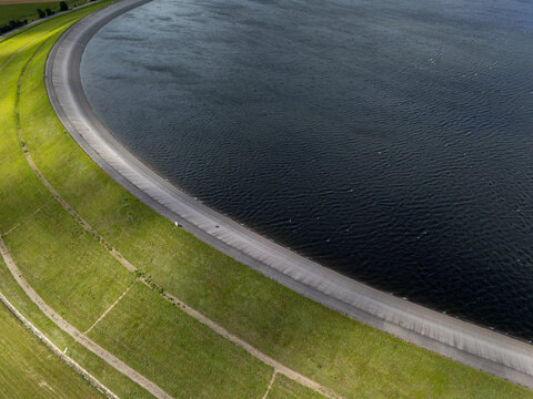 Aerial view of a large reservoir with a curved concrete dam embankment and lush green grassy slopes bordering the dark water in Poland.