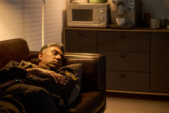 Middle aged security guard sleeping on sofa during break in office lounge, showing fatigue and overwork. Useful for workplace stress, shift work, burnout, staffing campaigns