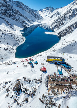 Aerial view of Portillo ski resort featuring the yellow hotel, parking lot, and deep blue Laguna del Inca surrounded by snow-covered Andes mountains in Portillo, Valparaiso, Chile.