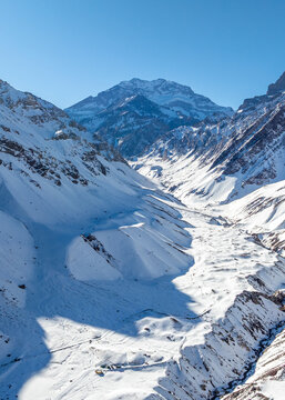 Aerial view of Aconcagua mountain peak rising above a vast snow-covered valley with rugged slopes and deep shadows under a clear blue sky, Las Heras Department, Mendoza Province, Argentina.