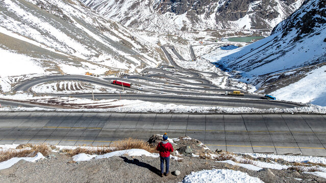 Aerial view of the winding Los Caracoles mountain road in the Andes with snow-covered peaks, trucks, and a person in a red jacket standing in the foreground in Portillo, Valparaiso, Chile.