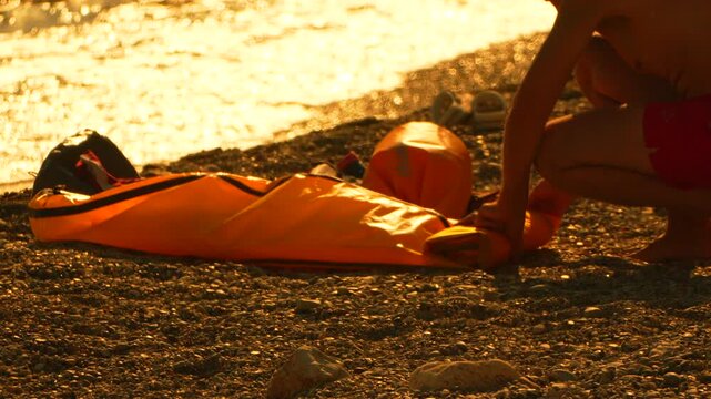 Orange drybag preparation occupies a swimmer on a pebbly shoreline during sunset. Medium side angle shot.