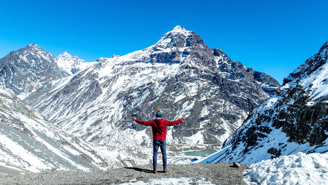 Aerial view of a person in a red jacket standing with arms outstretched facing the snow-capped Andes mountains under a clear blue sky in Portillo, Valparaiso, Chile.