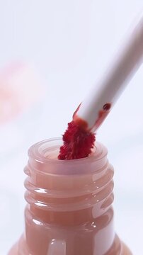 Red lipstick brush emerges from pink bottle in vertical front close up. Beauty cosmetic makeup applicator and packaging concept on blurred white background.