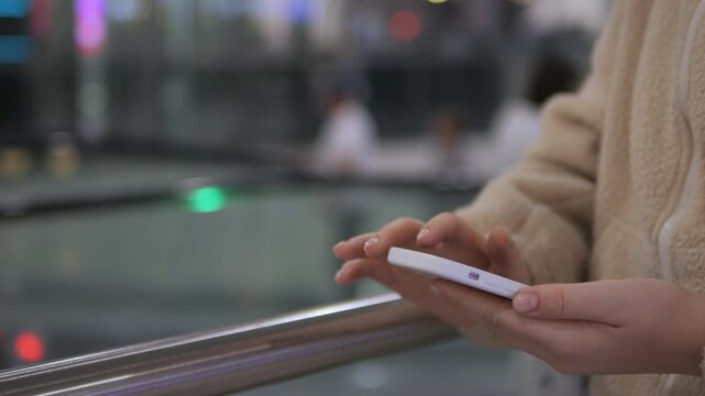 Person holds smartphone while standing on moving walkway. Fingers tap screen, focused on device. Background shows blurred travelers and glass railing. Ideal for travel, technology