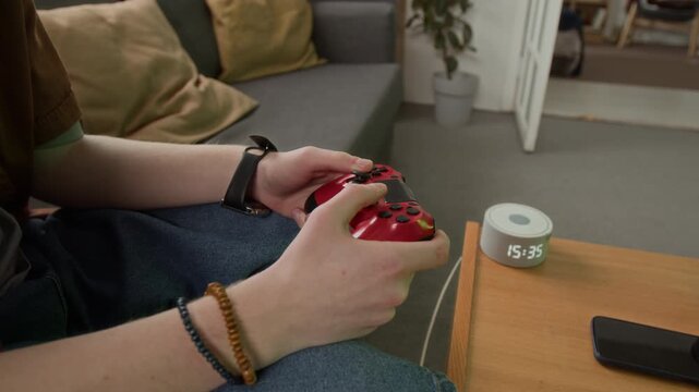 Hands of unrecognizable teenager playing video game with red controller, sitting on sofa near table with digital clock. Clip suitable for gaming, entertainment, or youth lifestyle marketing campaigns
