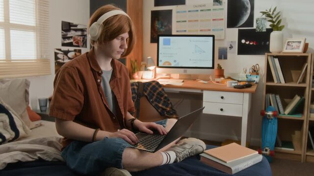 Caucasian teenager wearing headphones sitting on bed and typing on laptop while studying in bedroom. Video is ideal for online education, e-learning platforms, and student lifestyle marketing