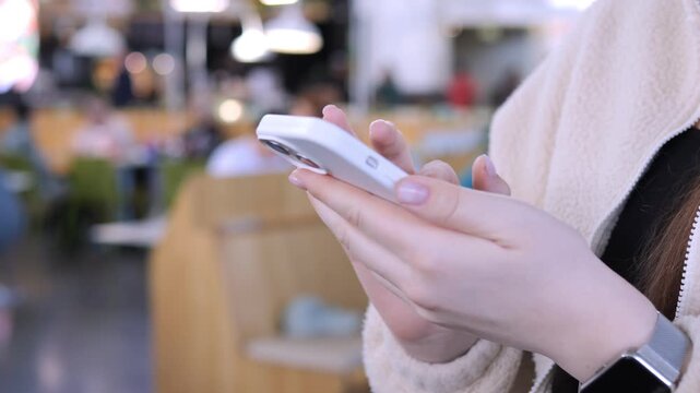 Person holds a smartphone while walking in a cafe. Their hands focus on the screen. Background shows blurred cafe seating and lights. The person appears calm and engaged