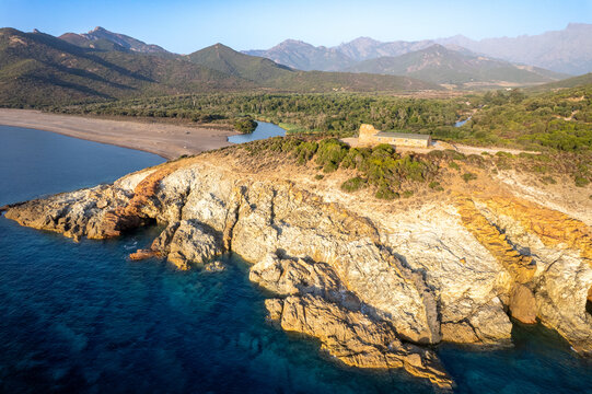 Aerial view of the historic Genoese tower overlooking the rugged coastline, turquoise sea, and the mouth of the Fango river with lush mountains in Galeria, Corsica, France.