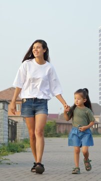 Loving young mother holding her little daughter's hand while walking together on a paved street in a residential neighborhood, enjoying a sunny day and creating precious family memories outdoors