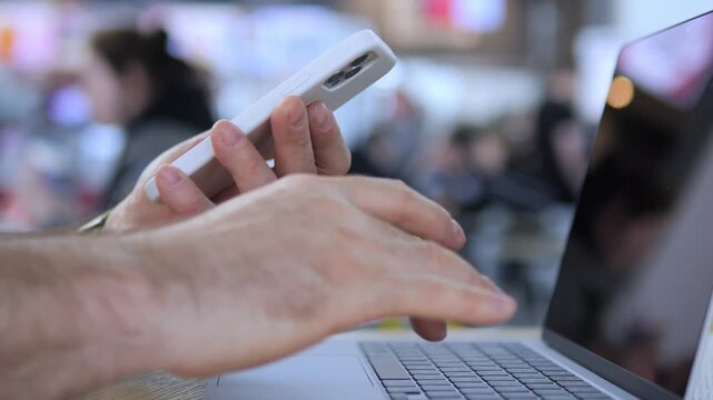 Close-up of hands using phone and laptop simultaneously. Fingers tap phone screen while typing on keyboard. Background shows blurred public space with yellow chairs. Person appears focused