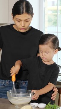 Adorable little child learning to cook while mixing batter in a bowl with a whisk, getting help from a caring mother. Happy family bonding and preparing breakfast together at home