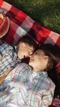 Happy mother and son laughing and tenderly touching noses while lying on a red checkered blanket during a summer picnic, sharing a joyful and loving moment together in the park
