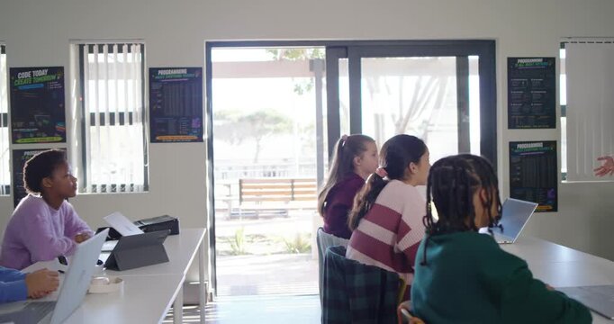Diverse teenage students turning to Indian teacher entering right and teaching coding on laptops