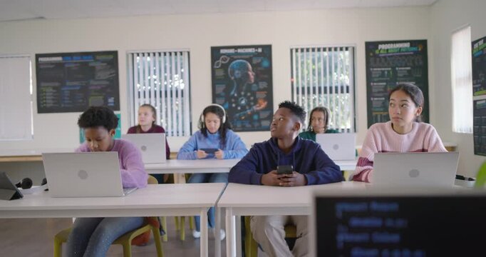 Diverse teen classroom watching teacher teaching at front, shifting focus from laptops to monitor