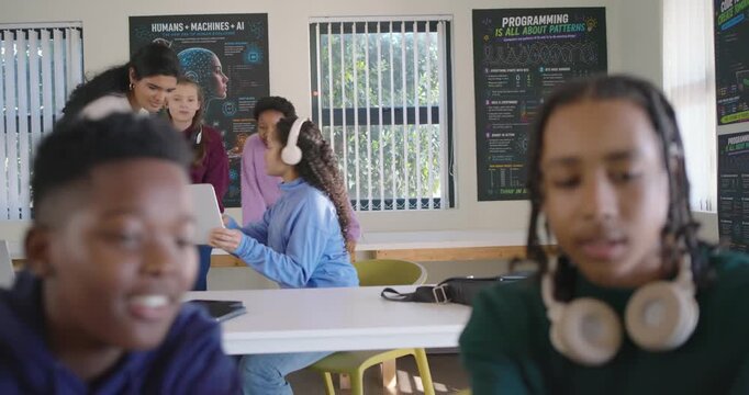 Two teenage male students leaning in at laptop in classroom, teacher approaching, working together