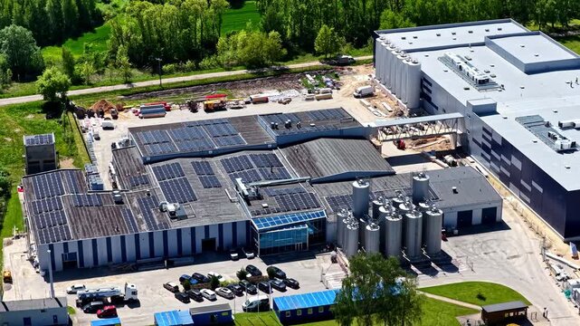 Riga, Latvia - 29 May 2025: Aerial view of industrial facility with solar panels on the roof, metal silos, and a parking lot surrounded by green fields.