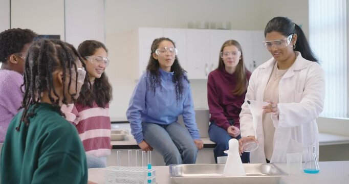 Indian teacher pouring in Erlenmeyer flask in lab as pupil handing vial causing foam, copy space