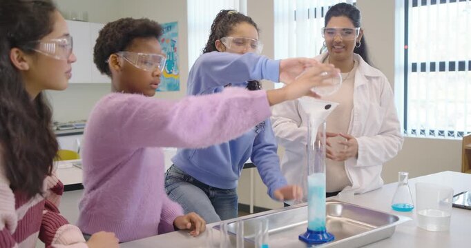 Indian instructor in lab coat, goggles guiding teen girls adding catalyst to cylinder in school lab