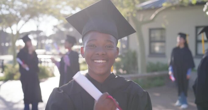 African American female grad laughing post-diploma on campus, holding cap and red ribbon diploma