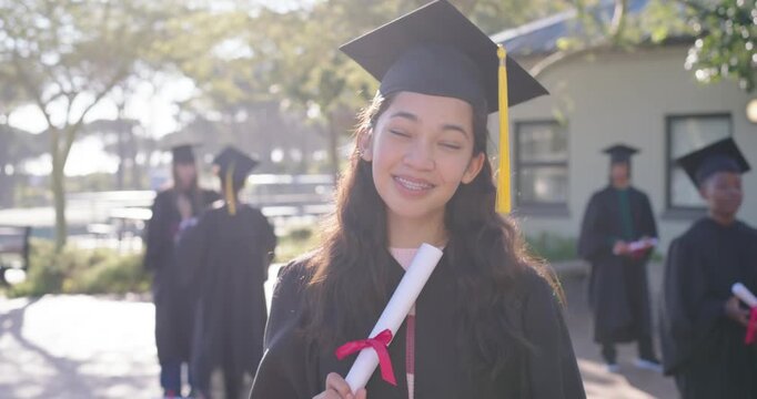 Indian woman in cap and gown extending red-ribbon diploma outward, posing on campus for graduation