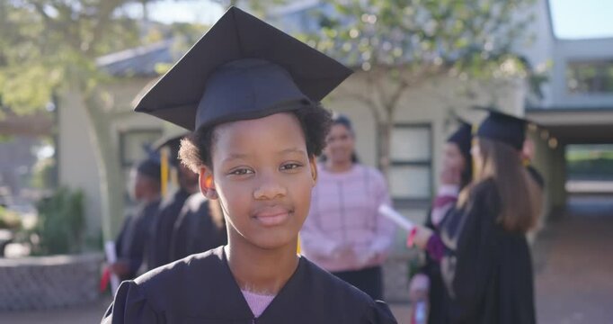 African-American gowned teen facing camera, smiling as camera zooming, grads holding diplomas quad