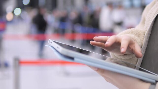Person holds tablet, taps screen. Blurred background shows busy public space. Red barrier tape visible. Hands in focus, fingers interact with device. Scene suggests travel or event setting