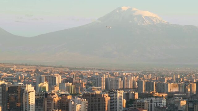 Scenic view of a large city in a high mountain valley and a descending passenger airplane against the city and a snow capped mountain. Passenger aircraft over Yerevan and Mount Ararat. Armenia.