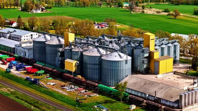 Riga, Latvia - 07 May 2025: Aerial view of a large grain storage facility with metal silos, a freight train on tracks, and surrounding green fields.