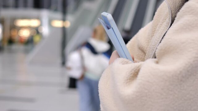 Person holds light blue tablet in airport terminal. Wears beige fleece jacket and smartwatch. Background shows blurred travelers and escalators. Focus stays on device and hands throughout frames