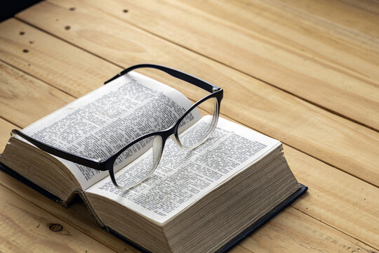 Black framed reading glasses resting on open pages of a thick English dictionary book lying on rustic wooden plank table, symbolizing nearsighted vision, literacy, education and learning concept