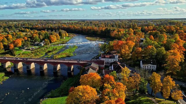 Kuldiga, Latvia - 06 November 2024: Aerial view of historic brick bridge spanning the Venta river surrounded by vibrant autumn foliage.