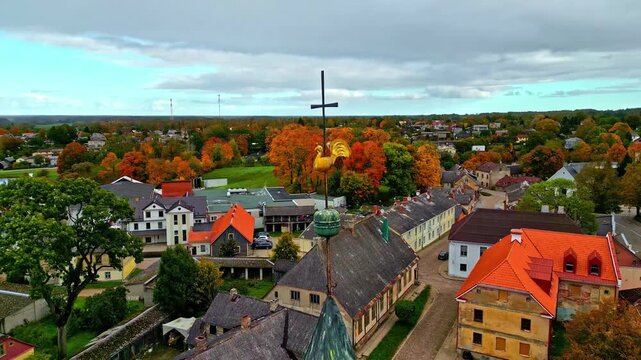 Riga, Latvia - 06 November 2024: Aerial view of the golden rooster weather vane atop a church spire overlooking historic buildings with red roofs and autumn trees.