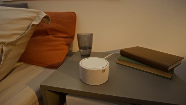 Smart speaker sitting on bedside table next to books and glass of water in bedroom. Footage is suitable for smart home technology and voice assistant marketing
