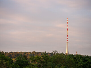 Obraz premium Cukrák television transmitter near Prague and Jíloviště illuminated by warm evening light. Tall broadcasting tower rising above forest under a colorful sunset sky.