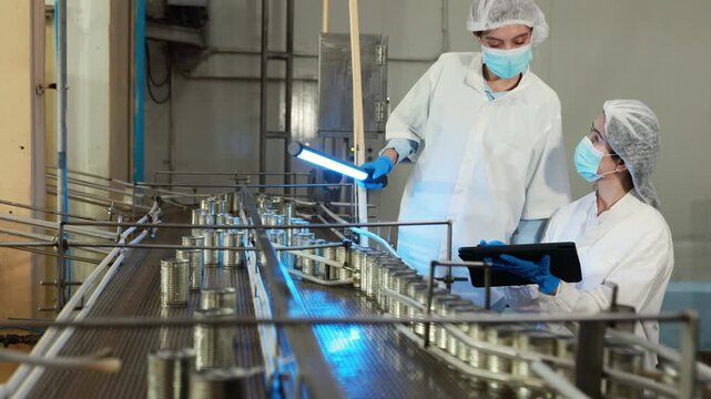 Team female researchers in white quality control suits holds RGB LED lights meticulously inspecting mackerel production line canning sauce filling conveyor belt industrial seafood processing plant.