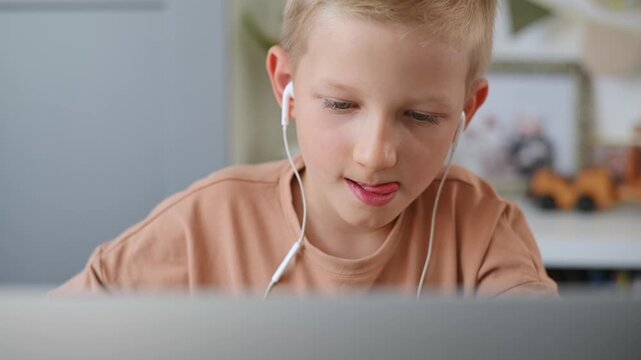 Schoolboy wearing headphones works intently on laptop. Small handsome boy stares at laptop screen and types on keyboard. Online learning and electronic lesson, modern technology. Slow motion video